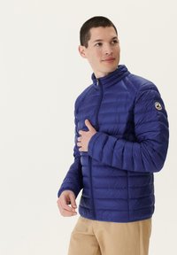 Chaqueta acolchada azul con cuello alto y cierre de cremallera, texturizada con costuras horizontales y un parche de logo en la manga izquierda.