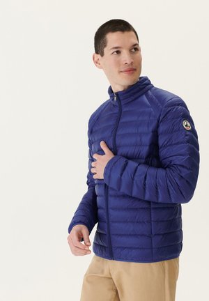 Veste bleue matelassée avec un col haut et une fermeture éclair, texturée avec des coutures horizontales, et un patch logo sur la manche gauche.