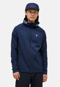 Chaqueta navy con cremallera y cuello alto, dos bolsillos laterales y detalles en logo blanco. La tela parece suave con un diseño ajustado.