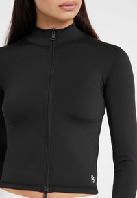 Giacca nera con zip e colletto alto, maniche lunghe e design aderente. Presenta una trama liscia e un logo ricamato vicino all'orlo.