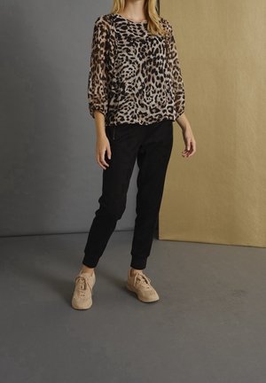 Vrouw draagt een doorschijnende blouse met luipaardprint, zwarte broek en beige sneakers, staand tegen een tweekleurige achtergrond in grijs en beige.