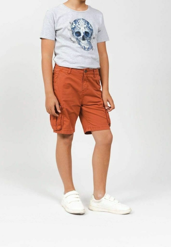 Enfant portant un t-shirt gris clair avec un motif de crâne floral bleu, un short cargo couleur rouille et des baskets blanches à velcro, debout sur un fond blanc.