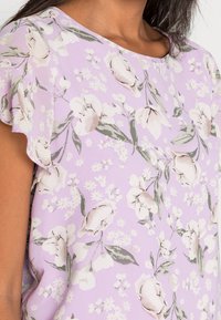 Top lilla con motivo floreale, maniche corte e volant. Presenta fiori bianchi e taupe con foglie verdi su uno sfondo morbido.