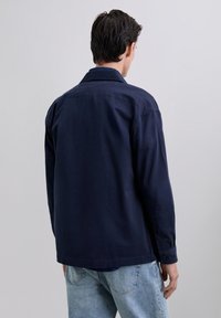 Hombre con cabello oscuro vestido con una camiseta de manga larga navy y jeans de color azul claro, de pie de espaldas contra un fondo claro y liso.