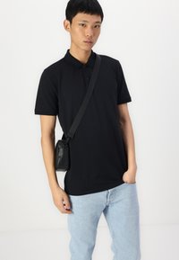 Camisa polo preta com uma abertura de três botões, combinada com jeans azul claro. Uma pequena mala cruzada preta pende do ombro.