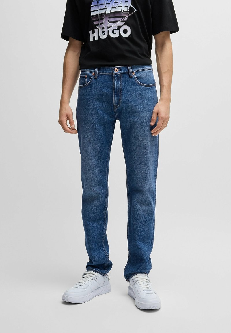 Blå denimjeans med rak passform, utrustade med en klassisk femfickedesign och subtila sömnadsdetaljer, ihop med vita sneakers.