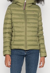 Doudoune verte olive avec capuche, couture horizontale et fermeture éclair à l'avant. Présente un petit logo sur le haut à gauche de la poitrine.