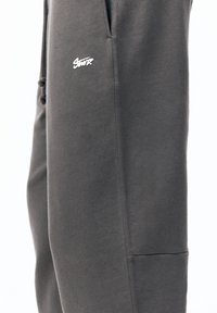 Grå joggingbukser lavet af blød stof med en glat tekstur. Der er sidelommer og et lille hvidt logo på venstre lår.