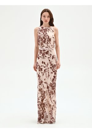 Jeune femme portant une robe beige sans manches, longue jusqu'au sol, avec un imprimé floral marron, debout devant un fond blanc uni.