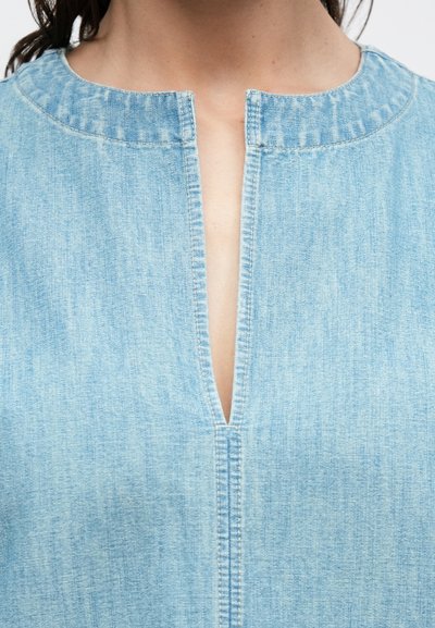 Κοντινό πλάνο ενός ανοιχτού μπλε ποδινού τζιν με στρογγυλή λαιμόκοψη και κεντρικό κάθετο άνοιγμα που αφήνει να φαίνεται το δέρμα.