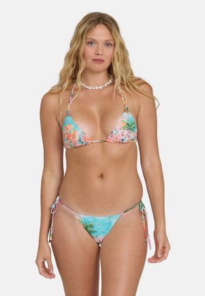 Donna bionda che indossa un bikini con stampa tropicale con palme e fiori, accessoriata con una collana di conchiglie bianche, su sfondo bianco.