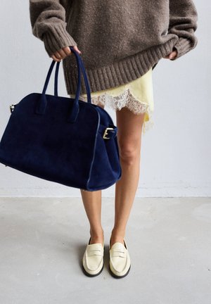 Persoon draagt beige loafers, gele jurk met kantafwerking, oversized bruine trui, houdt grote donkerblauwe handtas vast.