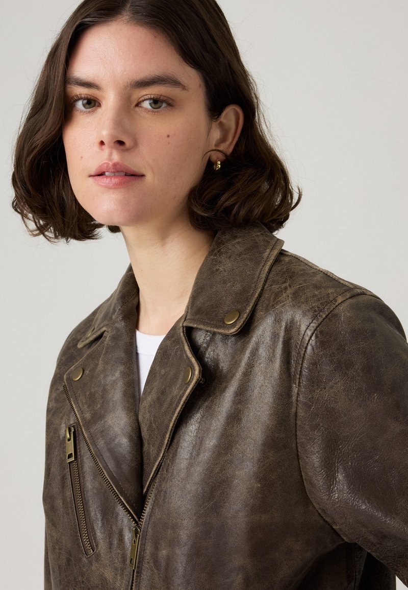 Veste de moto en cuir marron avec une finition texturée, épaulettes, fermeture éclair asymétrique et quincaillerie en laiton mate.