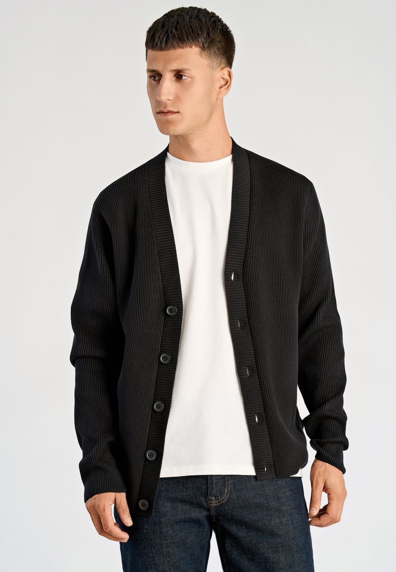 Czarny prążkowany sweter zapinany na guziki z głębokim dekoltem w serek, długimi rękawami oraz teksturowaną tkaniną, noszony na białej koszuli.