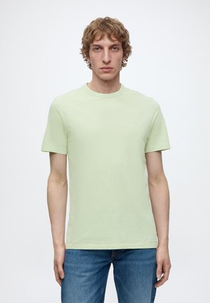 Jeune homme aux cheveux blonds bouclés portant un t-shirt vert clair à manches courtes et un jean bleu, debout devant un fond blanc uni.