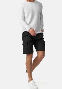 Haut blanc à manches longues avec une finition texturée, associé à un short cargo noir avec poches latérales. Le modèle porte des baskets blanches.