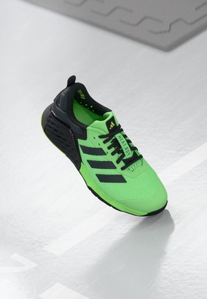 Chaussure de sport vert vif et noir avec des lacets noirs et trois bandes noires sur le côté, montrée flottant au-dessus d'un sol gris clair.
