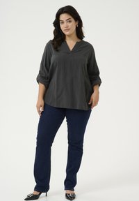 Blouse gris charbon avec un col en V et des manches roulées, associée à un jean bleu foncé et des chaussures à bout pointu noires et brillantes.