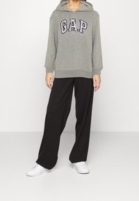 Sudadera con capucha gris con el logo "GAP", combinada con pantalones negros holgados y zapatillas blancas. La sudadera cuenta con un bolsillo tipo canguro y puños acanalados.