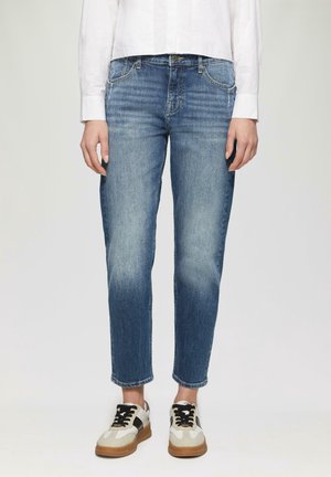 Person iført midt-højt blå denim jeans og hvide sneakers med sorte snørebånd, stående mod en ensartet lys baggrund.