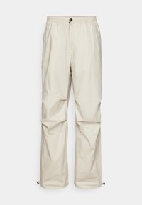Pantalones beige de cintura elástica con cordón ajustable, piernas rectas, costuras laterales y cierres de toggles en el dobladillo. Textura suave.