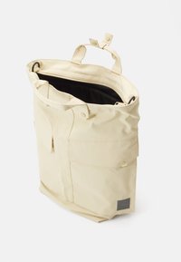 Beige taška přes rameno vyrobená z odolné tkaniny, s uzávěrem na zip, dvěma krátkými držadly a vnějšími kapsami pro dodatečné úložné prostory.