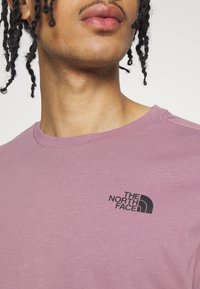 Světle fialové tričko s kulatým výstřihem vyrobené z bavlny, s černým logem The North Face na levé části hrudníku a hladkou texturou.