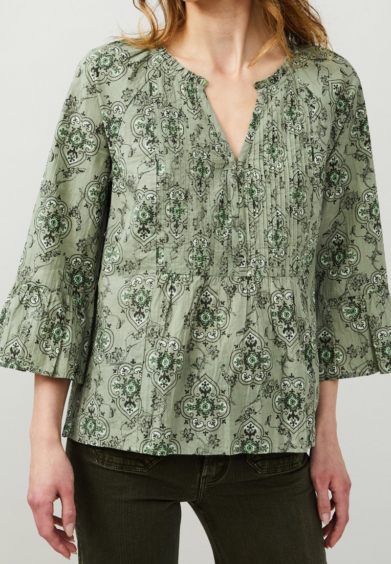 Grön mönstrad blus med V-ringning, veckad front och trekvartslånga trumpetärmar. Har intrikata blommönster och geometriska mönster.
