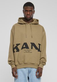 Felpa color khaki con grande stampa nera "KAN", cappuccio con cordino e tasca frontale. Presenta polsini e orlo a coste. Indossata sopra jeans azzurri chiari.