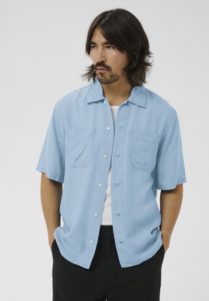 Mężczyzna o średniej długości ciemnych włosach, ubrany w jasnoniebieską koszulę na krótki rękaw zapinaną na guziki, na białej bluzce, z rękami w czarnych kieszeniach spodni.