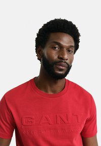 T-shirt in cotone rosso con logo "GANT" in rilievo e testo "AMERICAN SPORTSWEAR", colletto rotondo e maniche corte. Texture liscia.