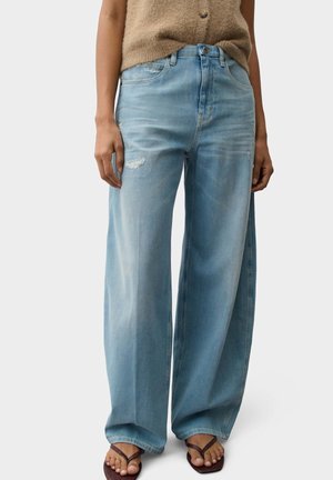 Person trägt hellblaue Jeans mit weitem Bein und kleinen Rissen, ein beiges ärmelloses Top und dunkelbraune Flip-Flops und steht vor einem einfarbigen Hintergrund.