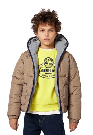 Veste matelassée de couleur beige avec un intérieur gris, présentant un col haut, des poignets élastiques et une texture matelassée. Portée par-dessus un sweat-shirt jaune vif.