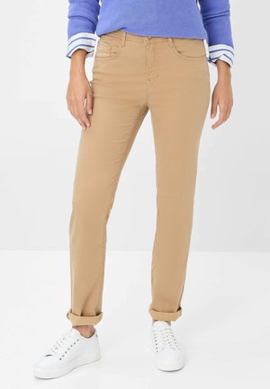 Beige Baumwollhosen mit geradem Bein, versehen mit Fronttaschen und umgeschlagenen Bündchen. Kombiniert mit weißen Sneakers und einem blauen Oberteil.