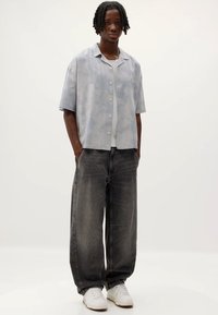 Chemise à manches courtes, rayée bleu clair avec des boutons, associée à un jean baggy gris foncé et des baskets blanches, se tenant contre un fond uni.