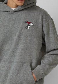 Grauer Hoodie aus weichem Material mit einem kleinen gestickten Snoopy-Design in einem roten Hut. Mit einer Fronttasche und gerippten Bündchen.