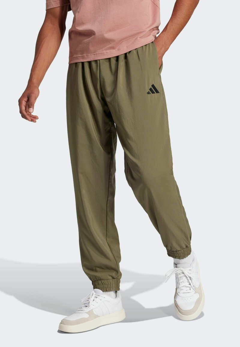 Olijfgroene trainingsbroek met elastische boorden en een zwart Adidas-logo aan de linkerzijde. Draagt met witte sneakers en een lichtroze shirt.
