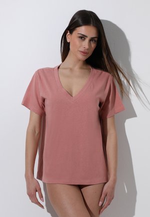 Donna con capelli lunghi e scuri che indossa una maglietta rosa polverosa con scollo a V e maniche corte, in piedi contro un muro bianco con ombra dietro di lei.