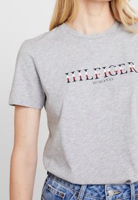 Camiseta de algodón gris de manga corta, con el logo en negrita "HILFIGER" en rojo y azul marino, y "MCMLXXXV" en una fuente serif clásica.