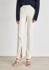 Crèmekleurige op maat gemaakte broeken met een hoge taille, zijsplitten en een gladde textuur, gecombineerd met beige sandalen met banden. Donkerblauwe trui erboven.