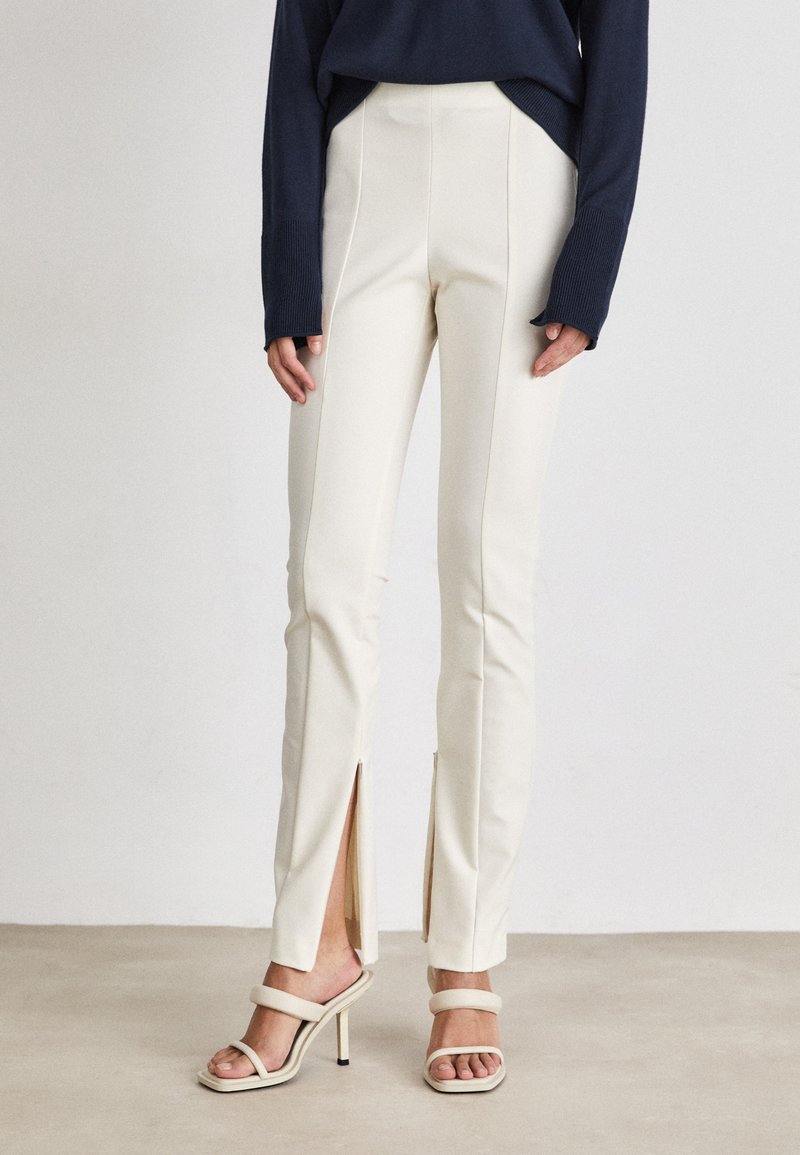 Crèmekleurige op maat gemaakte broeken met een hoge taille, zijsplitten en een gladde textuur, gecombineerd met beige sandalen met banden. Donkerblauwe trui erboven.