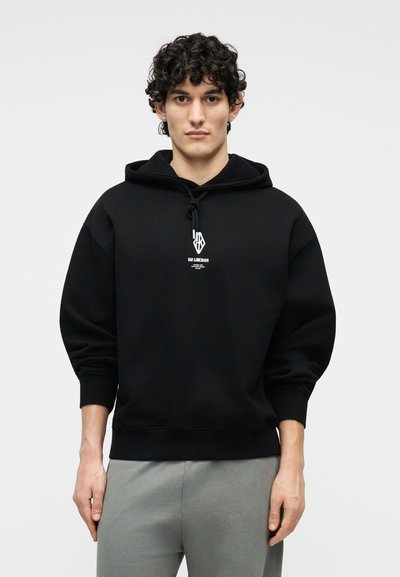 Νεαρός με σγουρά μαλλιά που φοράει μαύρο φούτερ με λευκό λογότυπο και γκρι παντελόνι, στέκεται μπροστά σε ένα απλό λευκό φόντο.