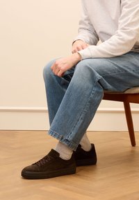 Braune Wildledersneaker mit dunklen Sohlen, kombiniert mit hellgrauen Socken. Locker sitzende blaue Jeans mit ausgefransten Säumen. Sitzt auf einem Holzstuhl.