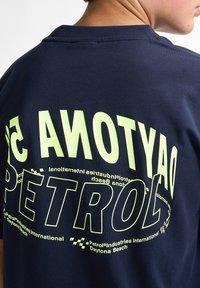 Camiseta azul marino con un gran texto verde claro que dice "DAYTONA" y "PETROL". Confeccionada en algodón, tiene una textura suave y un cuello tipo crew.