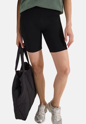 Kvinde iført sorte biker-shorts, sølvfarvede sneakers, holder en sort tote taske og en grøn top, der kun er synlig ved kanten.
