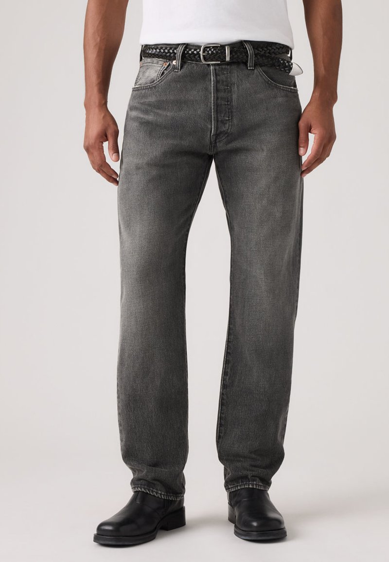 Άνδρας που φορά ξεθωριασμένο μαύρο τζιν με ίσια παντελόνια, μαύρη πλεκτή ζώνη, μαύρα μποτάκια και λευκό πουκάμισο που είναι μπαγιατισμένο μέσα, στέκεται μπροστά σε απλό φόντο.