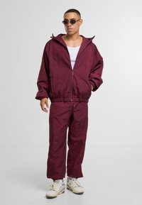 Bordowy, lekki dres z zapinaną na zamek kurtką, elastycznymi mankietami i dopasowanymi spodniami. Model nosi białą koszulkę na ramiączkach i okulary przeciwsłoneczne.