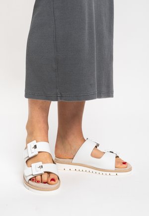 Hvide sandaler med to brede stropper og metalliske spænder, med en tekstureret sål og en glat overdel. Båret med en grå nederdel.