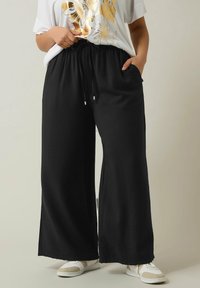 Zwarte wijde broek met een gestructureerde stof, voorzien van een elastische tailleband en zijzakken. Gepaard met een witte t-shirt en sneakers.