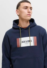 Tumesinine kapuutsiga sviiter, mille esipooles on logopatch, kus on boldis kirjas "JACK & JONES". Tõmbluku sulgemine roheliste aktsentidega ja kenguru tasku.